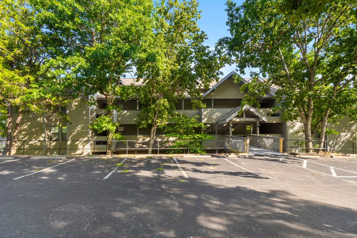 An exterior view of the building with ample parking spaces, shaded by mature trees and surrounded by landscaping. Guests are seen walking and socializing, demonstrating easy access and the welcoming environment.