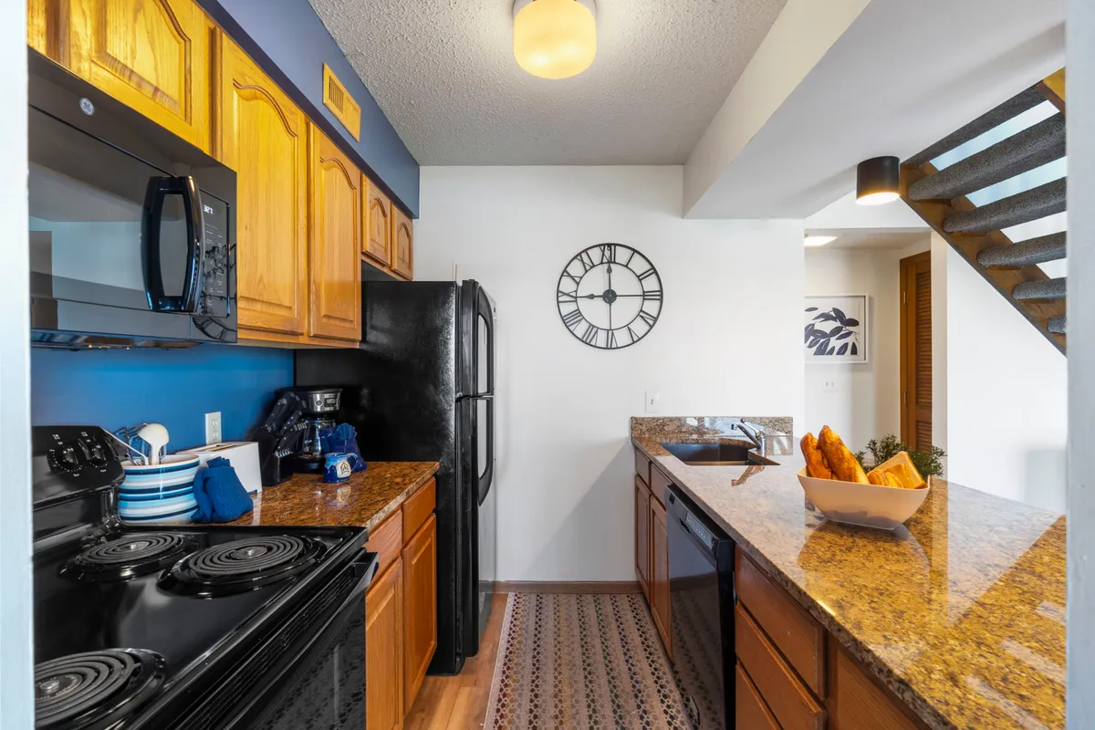 This image showcases a stylish and functional kitchen with granite countertops, stainless steel appliances, and warm wood cabinetry. A decorative clock adds a homey touch, while the open layout hints at easy interaction with the adjoining living areas.