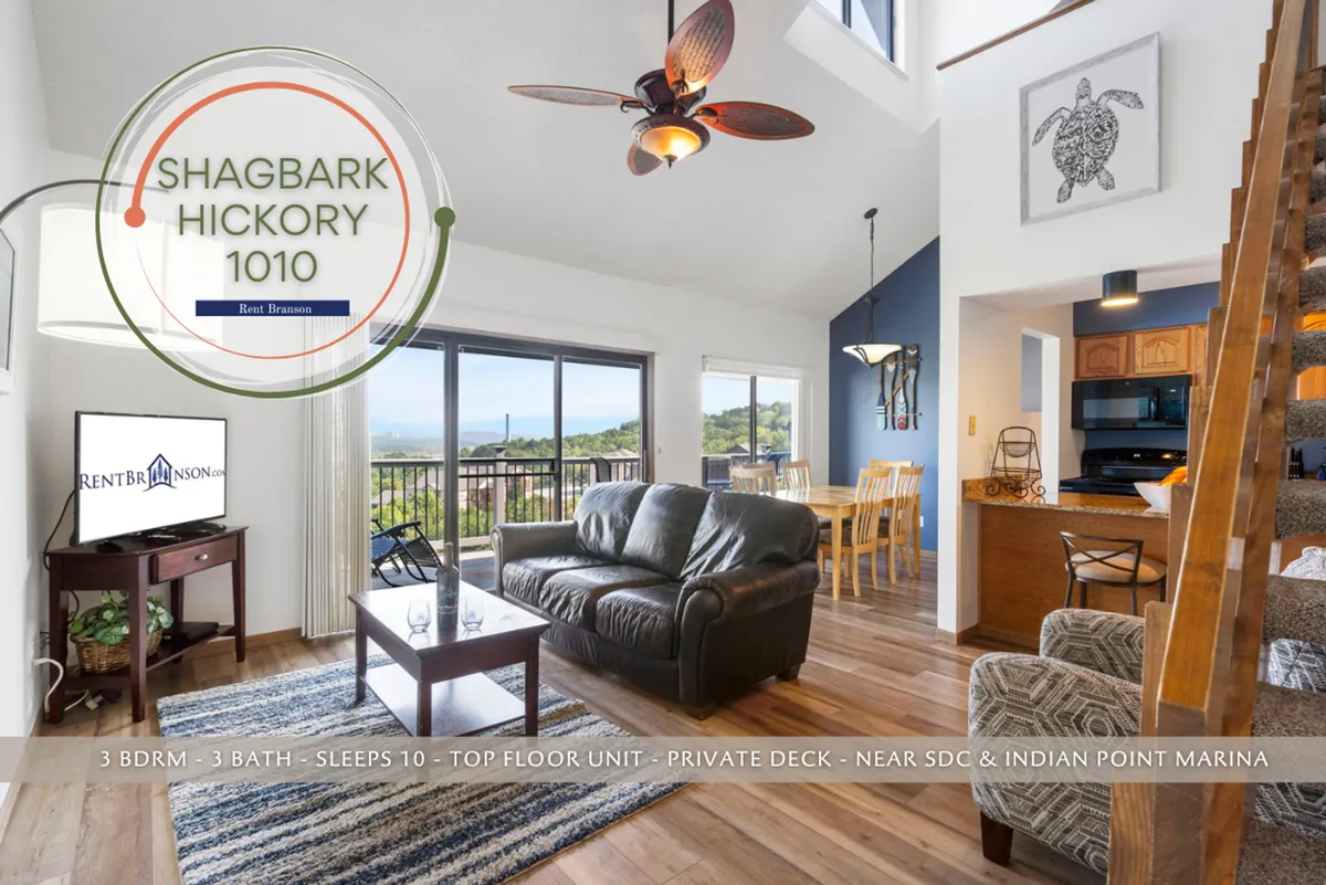 A bright, open-plan living area featuring large windows that frame a scenic view of the outdoors. The space includes a comfortable leather sofa, a dining table close to the windows, a wooden staircase, and tasteful wall art. Visible through the windows is a private deck accessible from the living area.