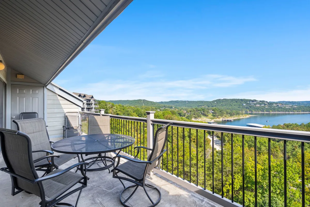A tranquil balcony setting featuring a table and chairs, with a sweeping view of the lake stretching out beyond the railing. The scene is calm, inviting guests to relax with a cup of coffee or take in the sunrise. The water is visible in the distance, and the overall atmosphere is serene and peaceful.