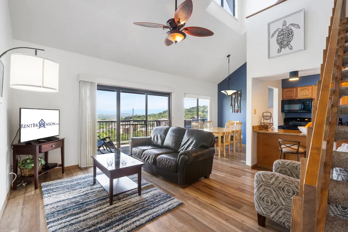 The image showcases a bright, inviting interior with a plush leather sofa, a spacious dining table for family meals, a television for cozy nights in, and expansive windows offering stunning, close-up lake views. The room feels open and airy, with natural light streaming in and a staircase leading to the top-floor vantage point. The scene evokes relaxation, togetherness, and breathtaking scenery right from the comfort of the living space.