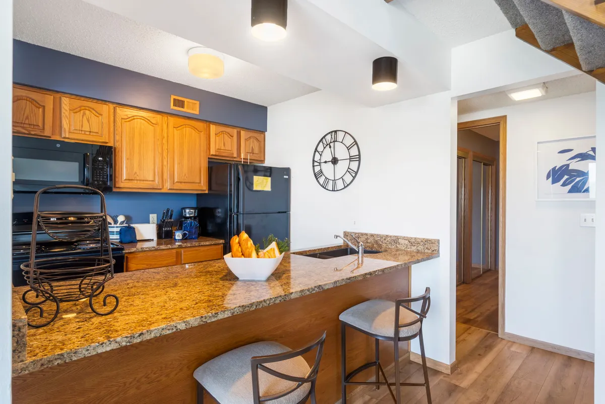 A bright kitchen scene featuring a granite countertop island, a fruit bowl, and a wall clock. The open design suggests ample space for meal prep and gathering, reinforcing the kitchen’s role as a social hub.