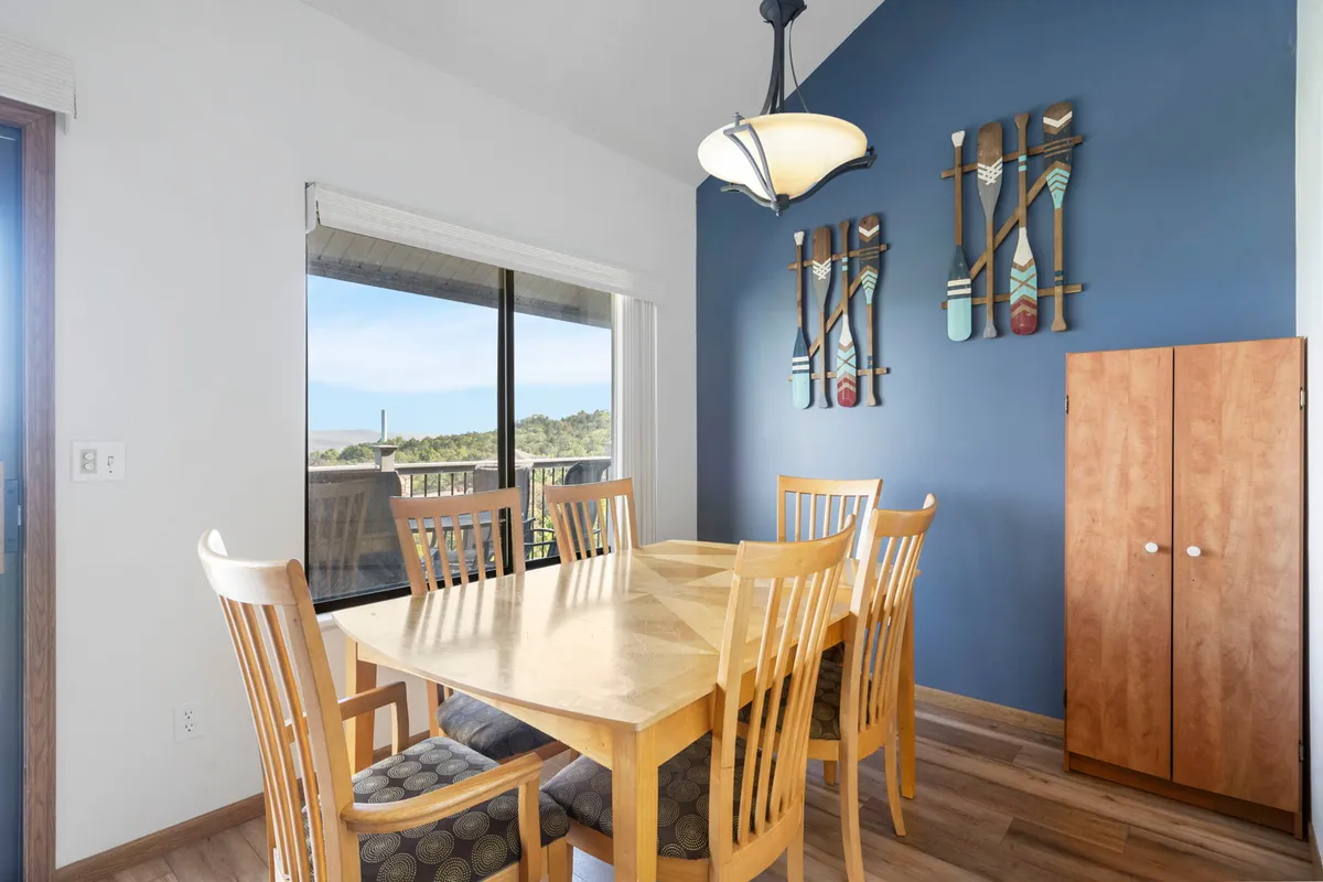 A welcoming dining area featuring a large wooden table, vibrant wall art, and a substantial window that allows natural light to pour in. The setup is ideal for group meals, games, and shared moments, all while providing a view of the outdoors.