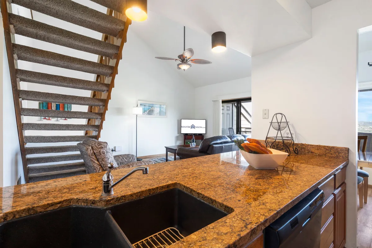 This view draws attention to the granite countertop stretching into an open living area, seamlessly connecting kitchen, dining, and relaxation spaces. The television and comfortable furnishings underline the kitchen’s integration into the heart of the home.